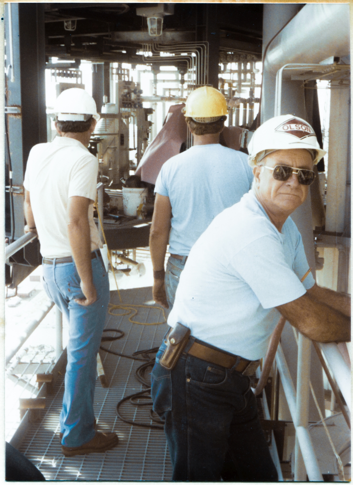 Image 096. At elevation 163'-9”, near the end of the OMBUU Access Catwalk on the Rotating Service Structure at Space Shuttle Launch Complex 39-B, Kennedy Space Center, Florida, Wade Ivey, owner of Ivey Steel, and Rink Chiles, Union Ironworker general foreman, stand side by side, discussing their immediate plans for working around whatever interference is at-the-moment preventing final positioning and bolt-up of the still-suspended OMBUU to the RSS. In the foreground, Olson Electric's general foreman, whose first name MIGHT have been Bill, and whose last name I cannot recall, turns and gives me a deeply-disapproving look as I hit the shutter release on my camera. Bill and I got along excellently, and we both helped each other on a regular and reliable basis when issues that intersected the worlds of steel erection and electrical installation came up, but, as with almost everyone else, he did NOT like the idea of a photograph being taken, of the facility in general, and which also included him in particular. It was a cultural barrier that I always had to deal with, and by this time I was long past worrying about it. I had a camera. I had a camera permit. And I fully intended to USE them. Period. Photo by James MacLaren.